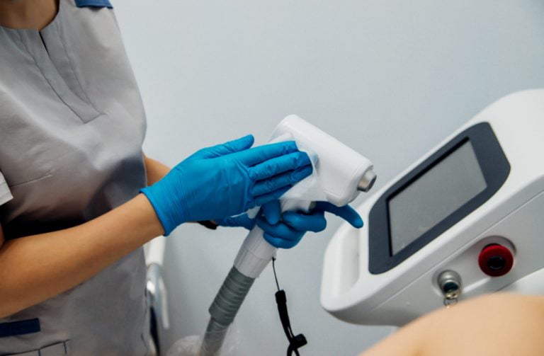 Women's hands in blue gloves hold laser hair removal equipment.